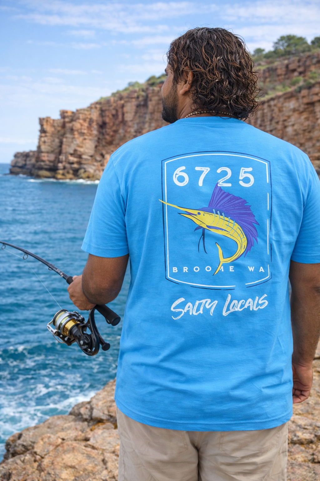Man wearing 6725 Sword Adult Tee while fishing by the ocean with rocky cliffs in the background.