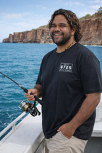 Man smiling while fishing in the ocean wearing a black 6725 Sword Adult Tee, enjoying a coastal lifestyle.