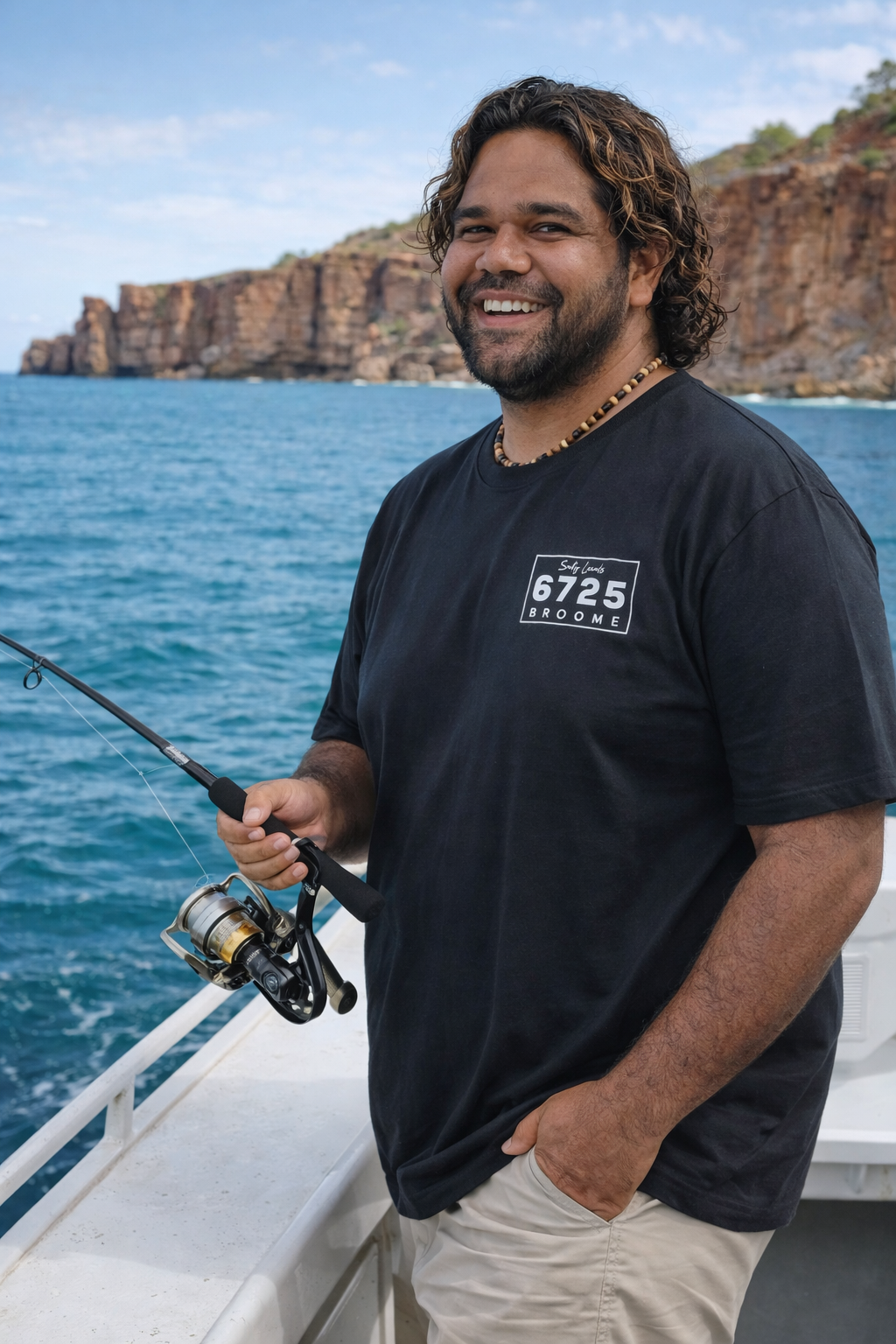 Man smiling while fishing in the ocean wearing a black 6725 Sword Adult Tee, enjoying a coastal lifestyle.