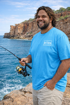 Man fishing by the ocean wearing the 6725 Sword Adult Tee in blue, showcasing coastal style and comfort.