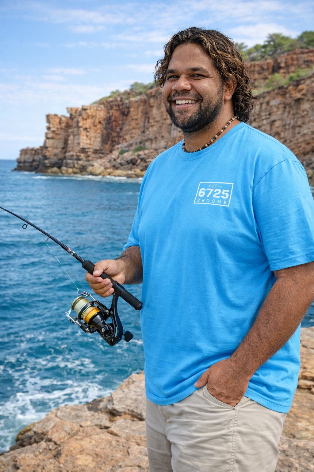 Man fishing by the ocean wearing the 6725 Sword Adult Tee in blue, showcasing coastal style and comfort.