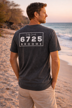 A man wears a Men's Stonewash T-Shirt - Black with 6725 Broome branding on the beach at sunset.